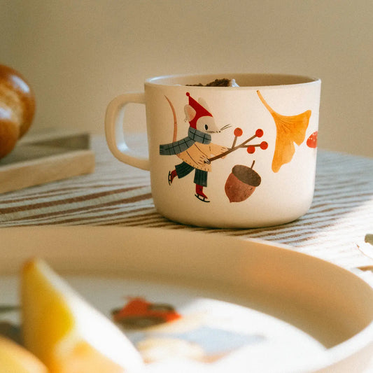 A cream-colored ceramic mug with a whimsical illustration of a mouse in a red hat and gray scarf holding a branch with red berries, alongside an acorn and orange ginkgo leaves. The mug is set on a striped table runner with a warm, softly blurred background.