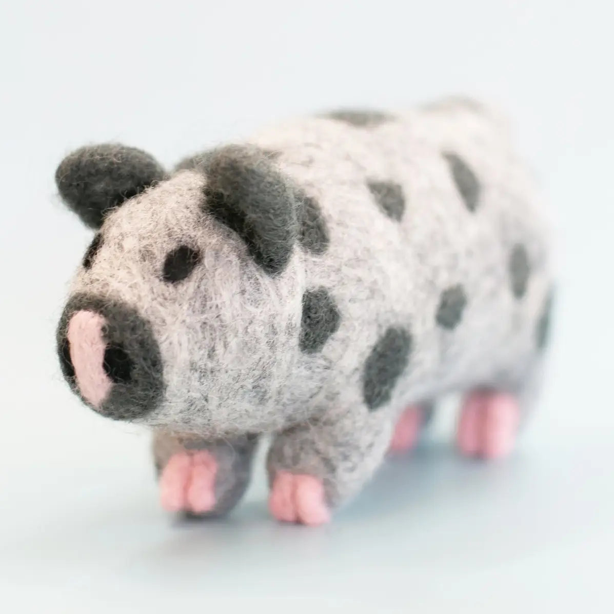 A small felted wool pig figurine with a gray and white spotted body, dark gray ears and snout, and pink feet and nose, photographed on a clean light background.