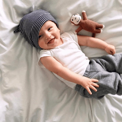 Baby lying on a bed wearing a striped hat and holding a monkey plush toy.