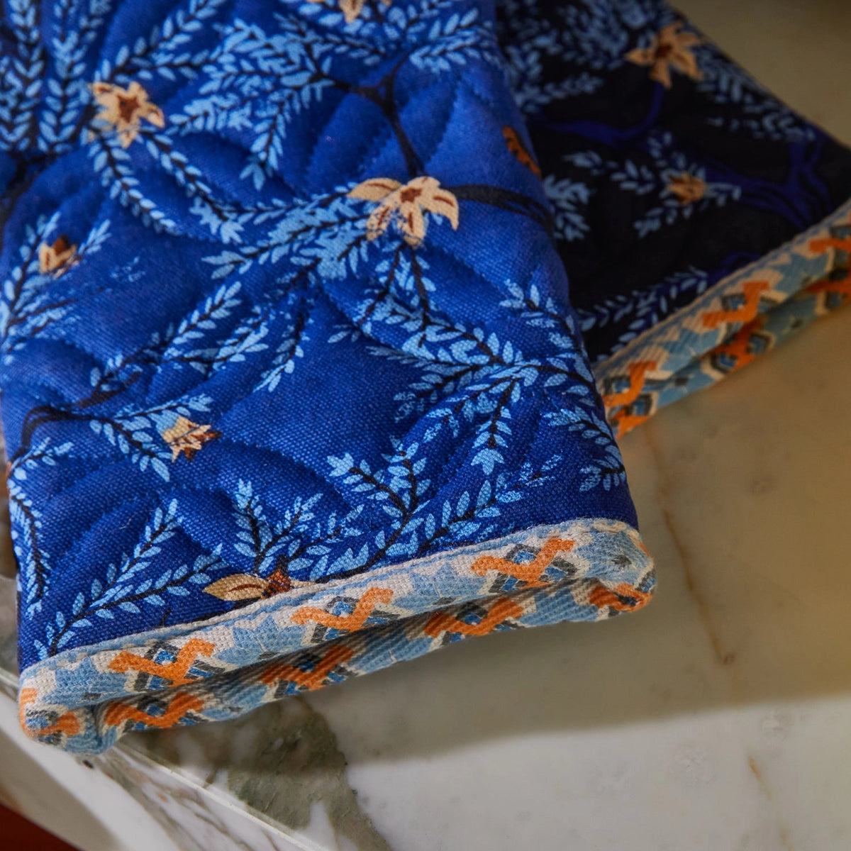 A close-up of a folded quilted textile, likely a blanket or quilt, featuring a blue floral and leaf pattern with orange and beige accents, resting on a light marble surface.