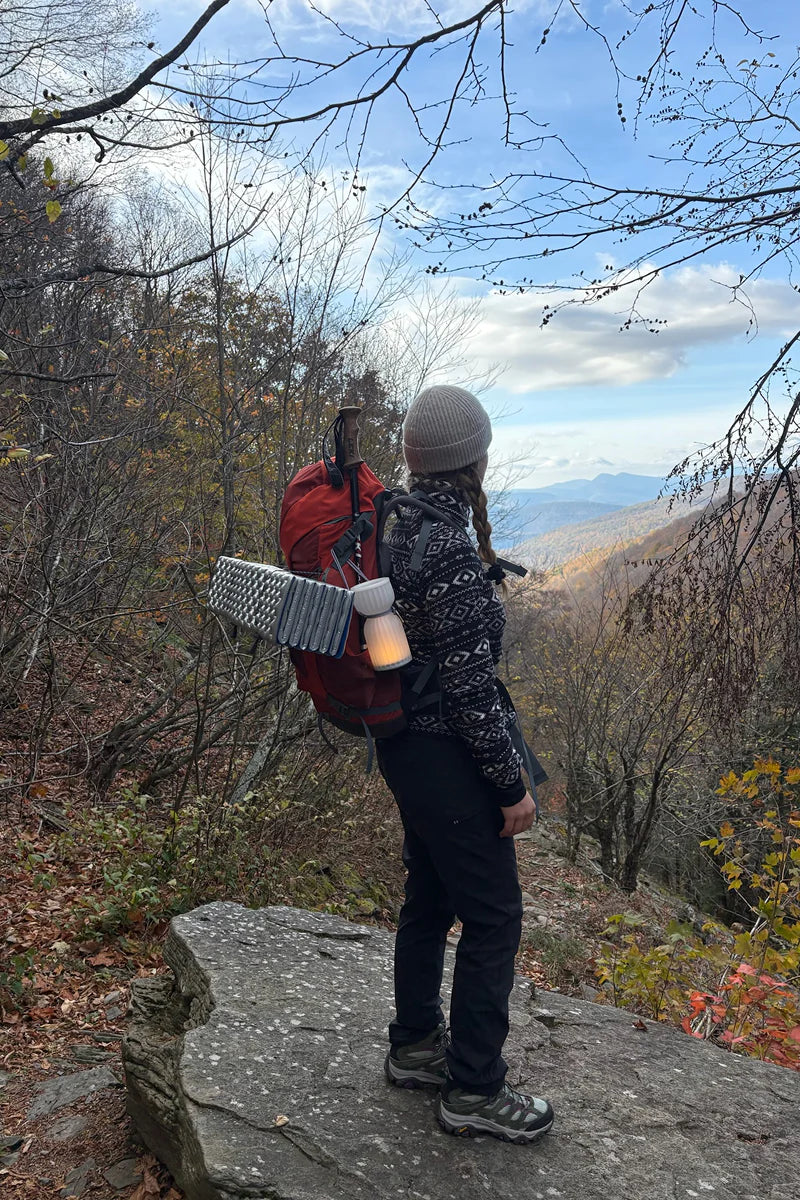Person with a red backpack and patterned jacket standing on a rocky outcrop in a forest with mountains in the background.