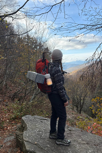 Person with a red backpack and patterned jacket standing on a rocky outcrop in a forest with mountains in the background.