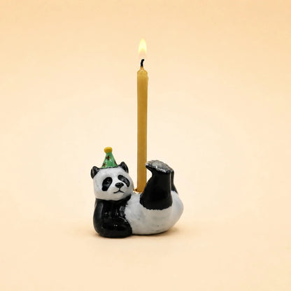 A ceramic panda candle holder with a black and white painted panda sitting on its back holding a single lit yellow candle. The panda wears a small green party hat.