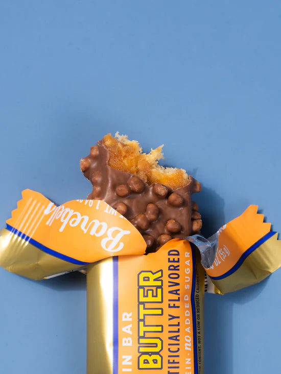 A partially unwrapped chocolate butter bar with a bite taken out, showing the chocolate-covered cookie filling and nut coating, set against a solid blue background.