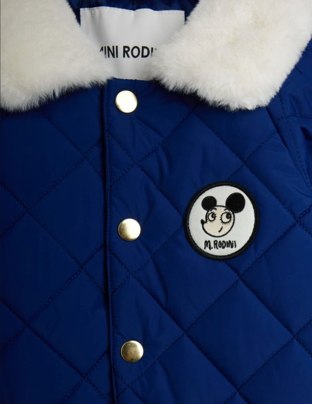 Close-up of a royal blue quilted puffer jacket with a white faux-fur collar, gold-tone snap buttons, and a circular black-and-white patch featuring a cartoon face and the text 'MINI RODI'.