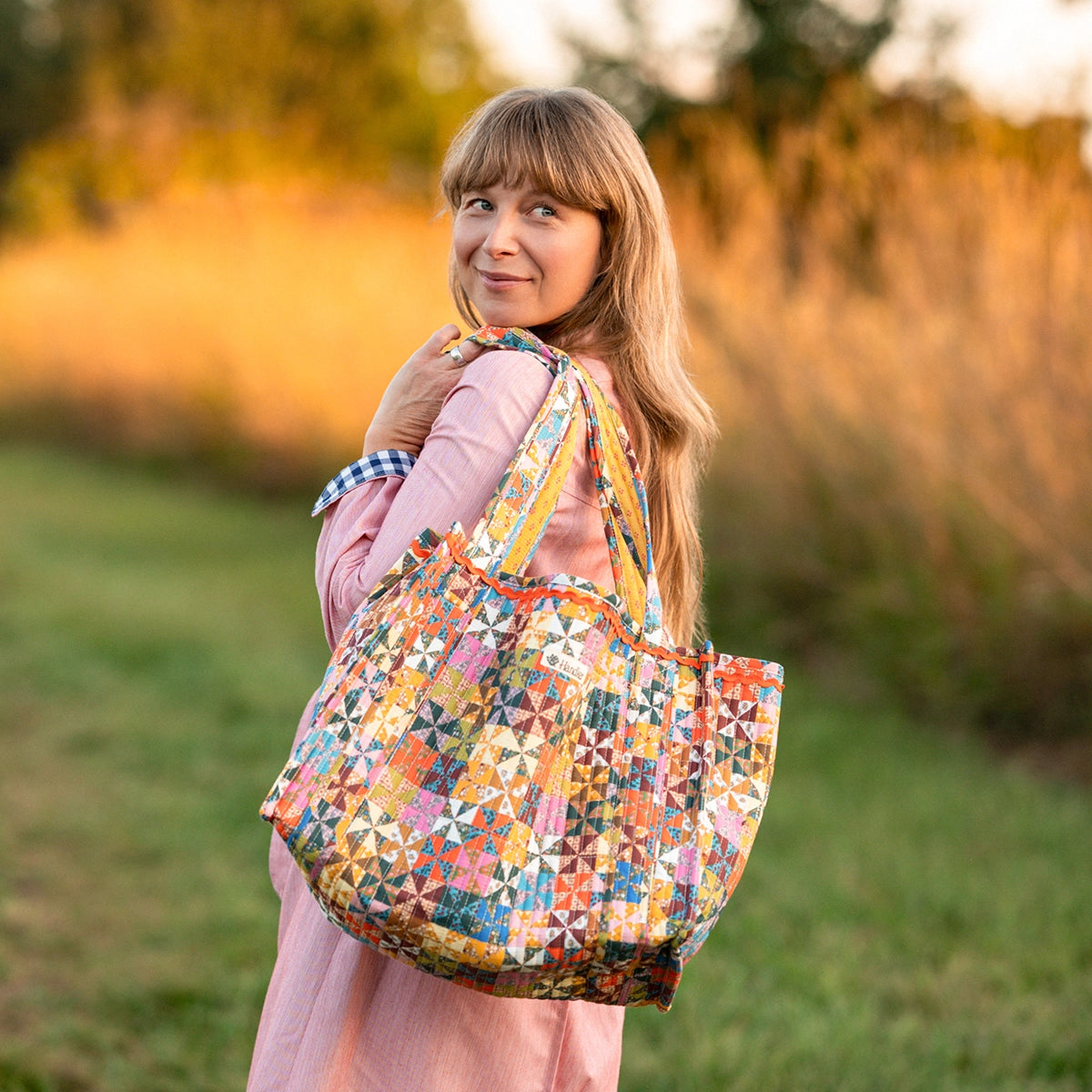 A woman in a pink dress carrying a large quilted patchwork tote bag outdoors in a grassy field at sunset.