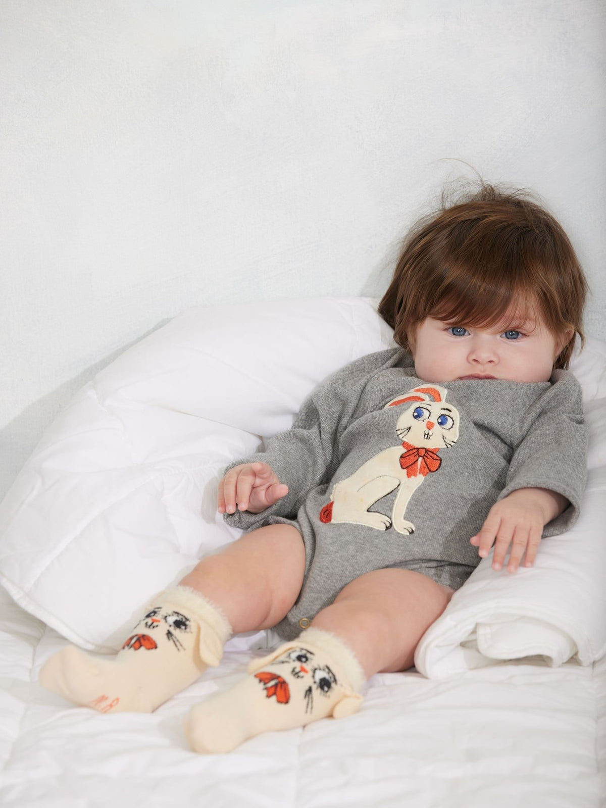 A baby lying on a white quilted bed, wearing a gray long-sleeve bodysuit with a white bunny graphic and cream-colored socks with a rabbit face design.
