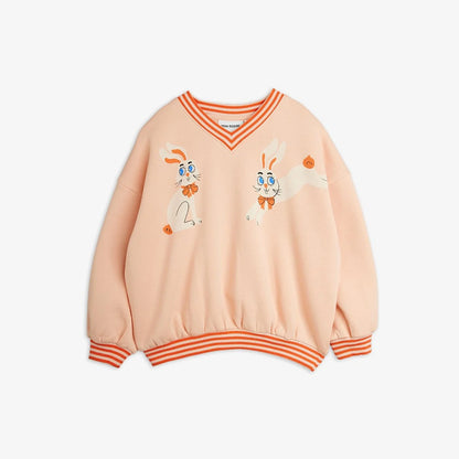 A light peach V-neck sweatshirt with orange and white striped ribbing on the collar, cuffs, and hem. The front features two cartoon bunnies wearing orange bowties and small orange pumpkins, set against a plain light gray background.