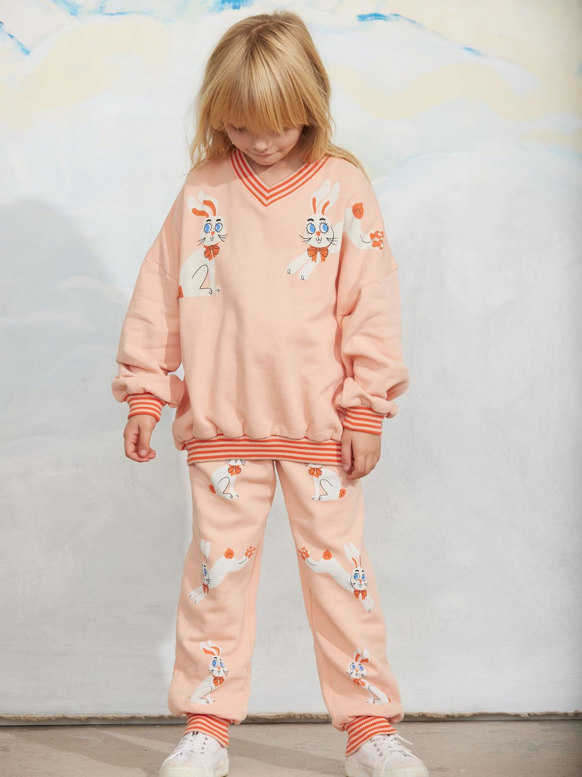 A young girl with blonde hair wearing a peach-colored bunny-themed sweatshirt and matching sweatpants with orange striped trim, standing against a textured light gray wall.