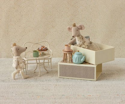 Miniature mouse figures with a tea set on a textured surface with a patterned wall background.