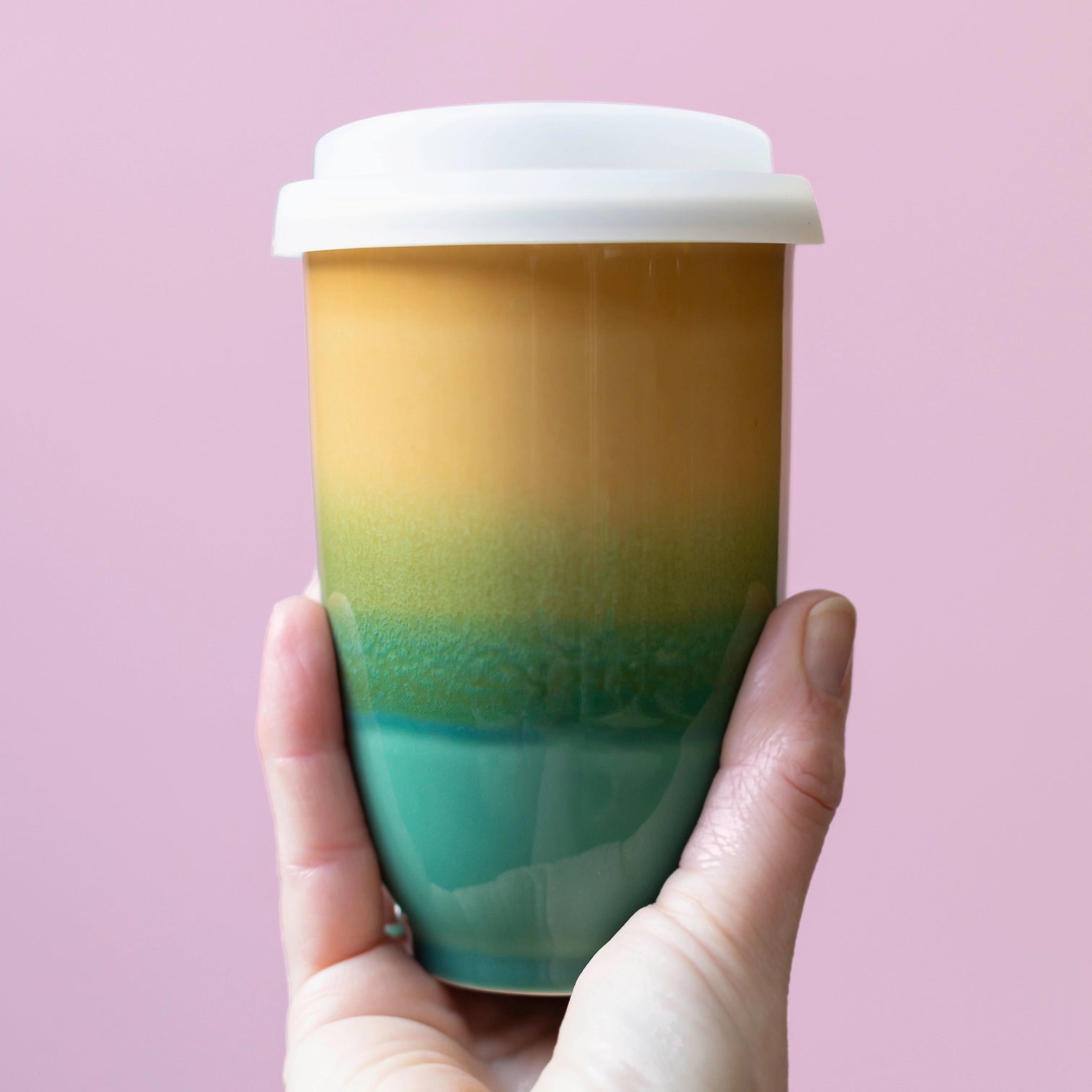 Hand holding ceramic, green and yellow travel mug on a pink background
