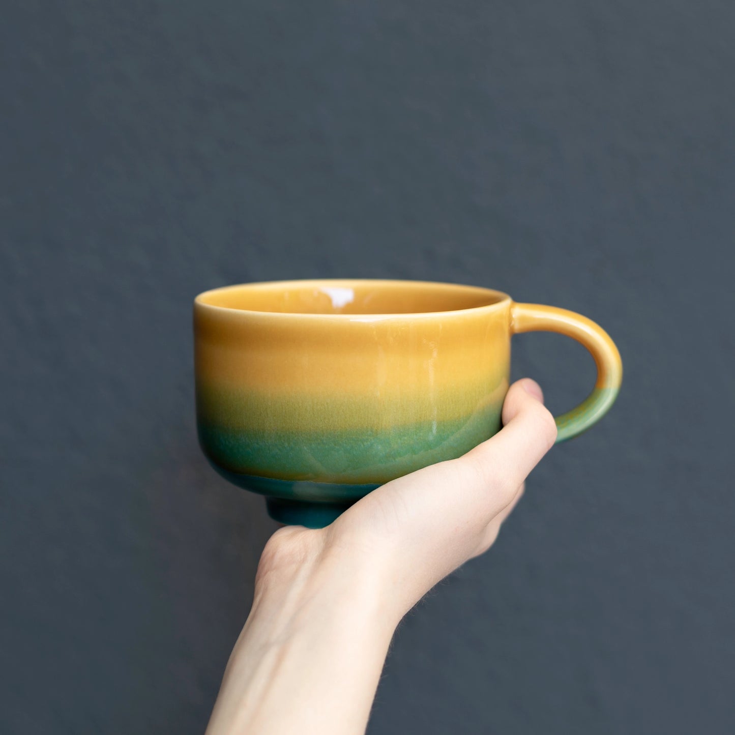A ceramic cup with a yellow-to-teal gradient glaze, held by a person's hand against a dark gray background.