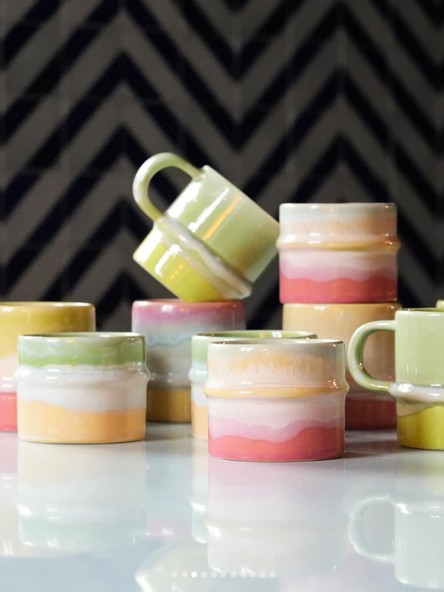 A group of small ceramic mugs with colorful horizontal stripes in pastel shades of pink, yellow, green, and orange, arranged on a glossy white surface with a dark chevron-patterned background.