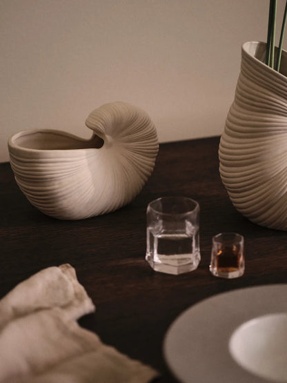A styled tabletop scene with two ribbed ceramic vessels in warm off-white tones, one shallow bowl-like form and one taller vase-like form, alongside clear drinking glasses and a folded neutral cloth on a dark wooden surface.