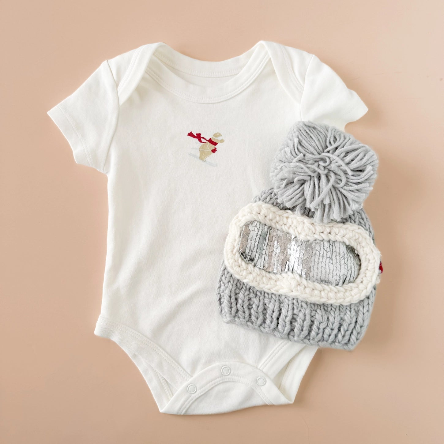 White baby onesie with a gray knit hat and goggles on a beige background