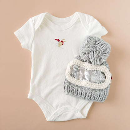 White baby onesie with a gray knit hat and goggles on a beige background