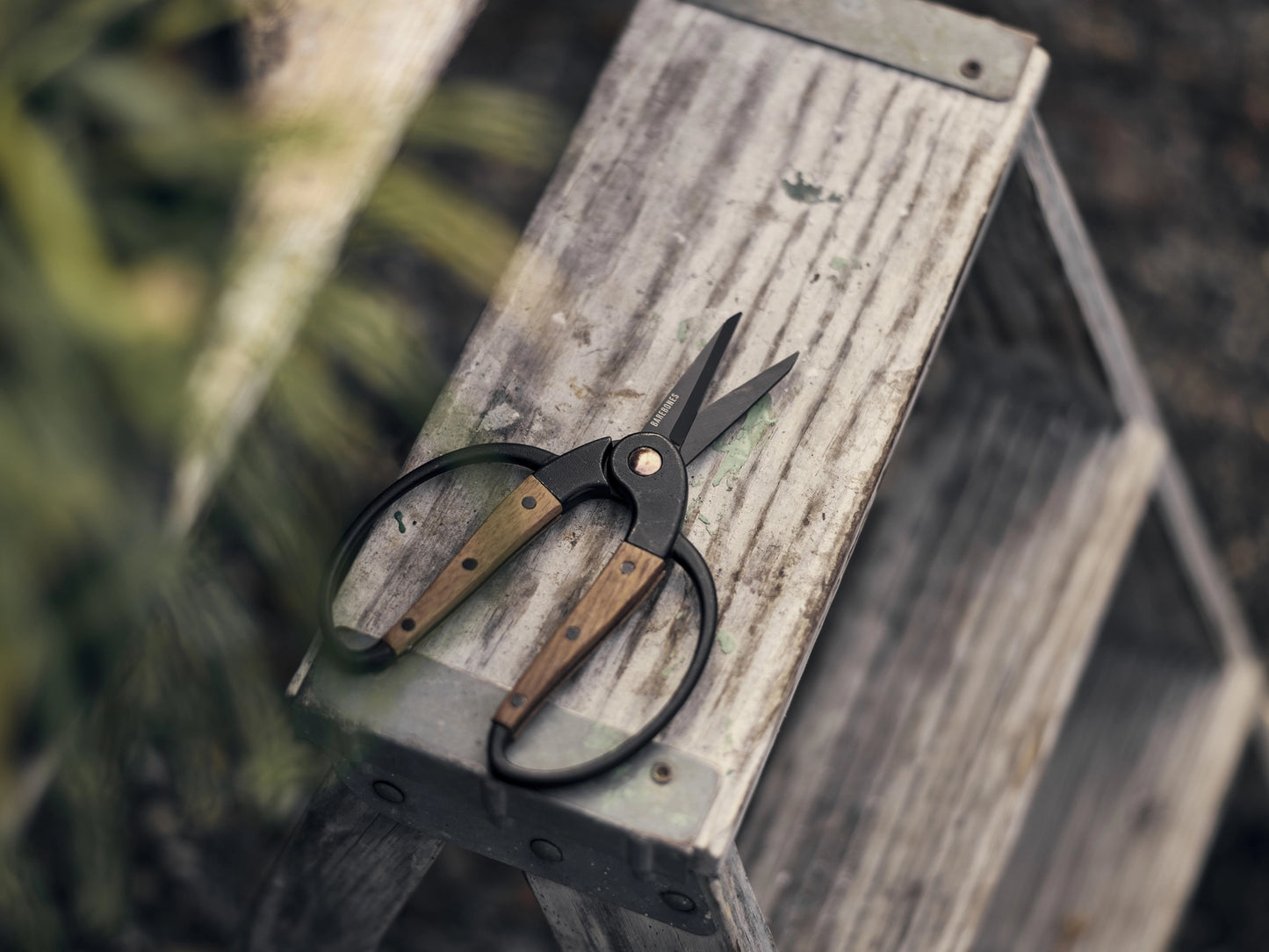 A pair of pruning shears with black metal blades and light brown wooden handles resting on a weathered gray wooden step stool.