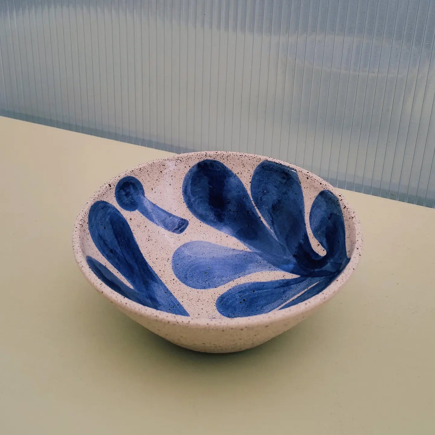 A handmade ceramic bowl with a speckled beige exterior and a blue hand-painted pattern inside, set on a light beige surface with a ribbed translucent window pane in the background.