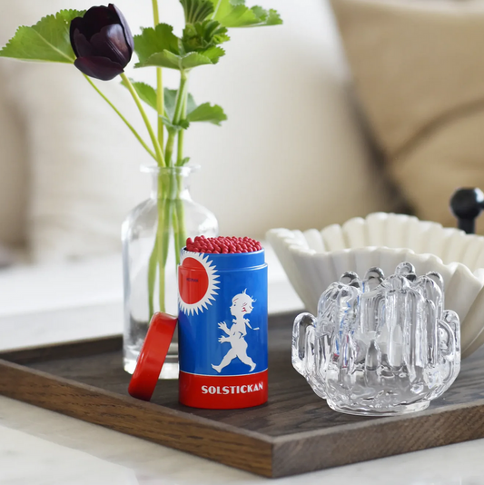Solestickan canister with red lid on a wooden tray with a glass mug and vase in the background.