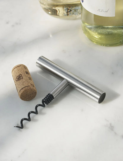 Silver corkscrew and cork on a marble surface with wine bottles in the background.