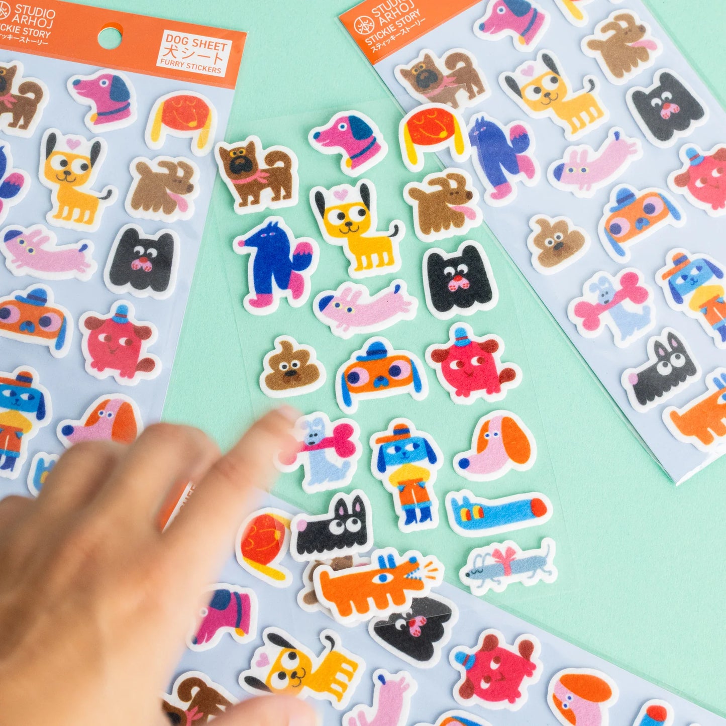 Three sheets of colorful cartoon dog stickers on a mint green background, with a person's hand reaching in to pick up one of the stickers.