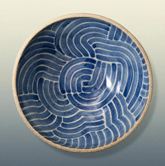 A round ceramic bowl with a blue and white wavy pattern inside and a textured beige rim, shown against a neutral gradient background.