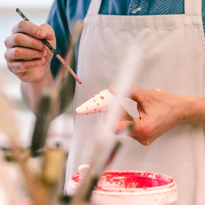 Person painting a ceramic item with a brush and pink paint, wearing an apron.