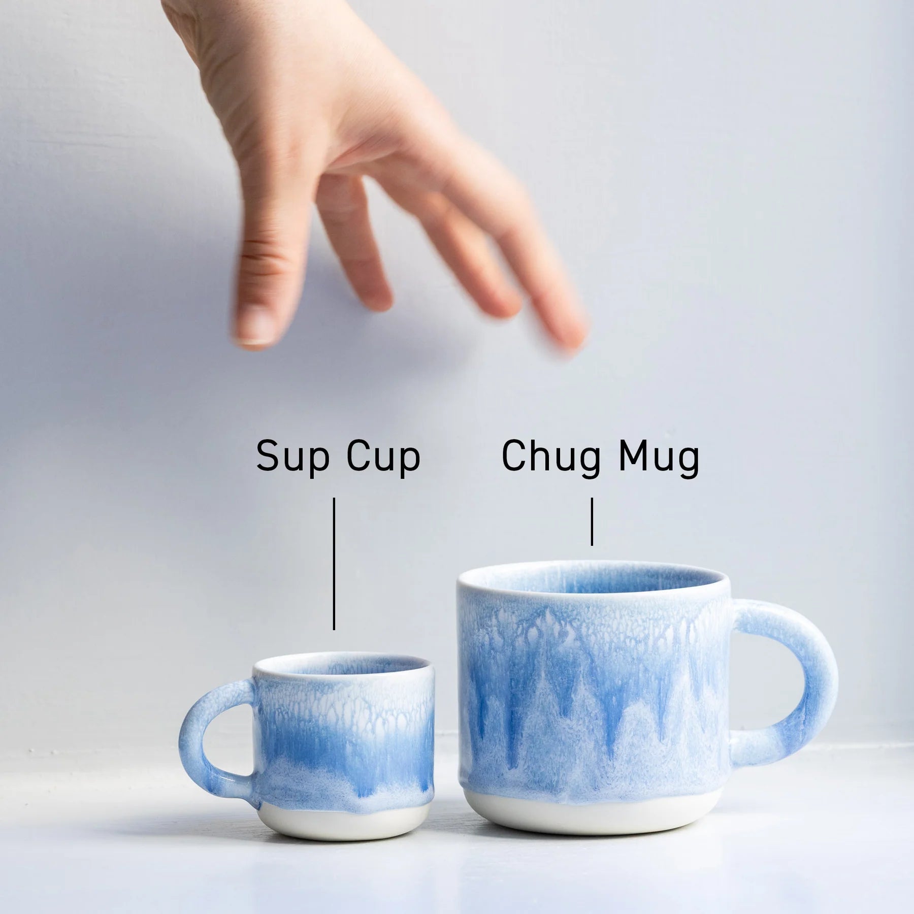 Two blue ceramic mugs labeled 'Sup Cup' and 'Chug Mug' with a hand reaching towards them.