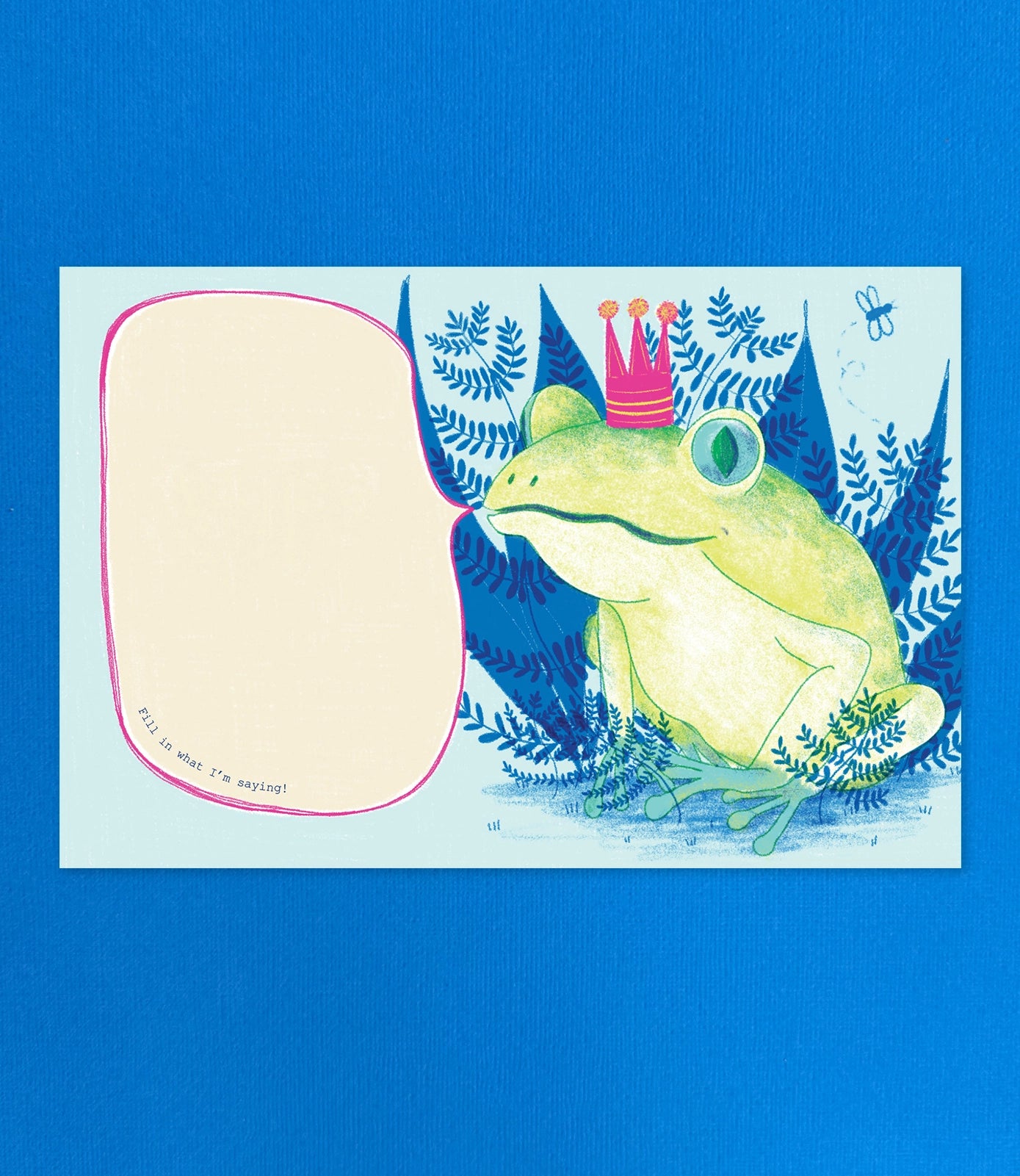 A square printed card or art print featuring a whimsical illustration of a green frog wearing a pink crown, surrounded by blue fern-like foliage, with a large blank beige speech-bubble shape on the left. The print is photographed against a solid bright blue background.