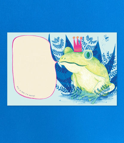 A square printed card or art print featuring a whimsical illustration of a green frog wearing a pink crown, surrounded by blue fern-like foliage, with a large blank beige speech-bubble shape on the left. The print is photographed against a solid bright blue background.
