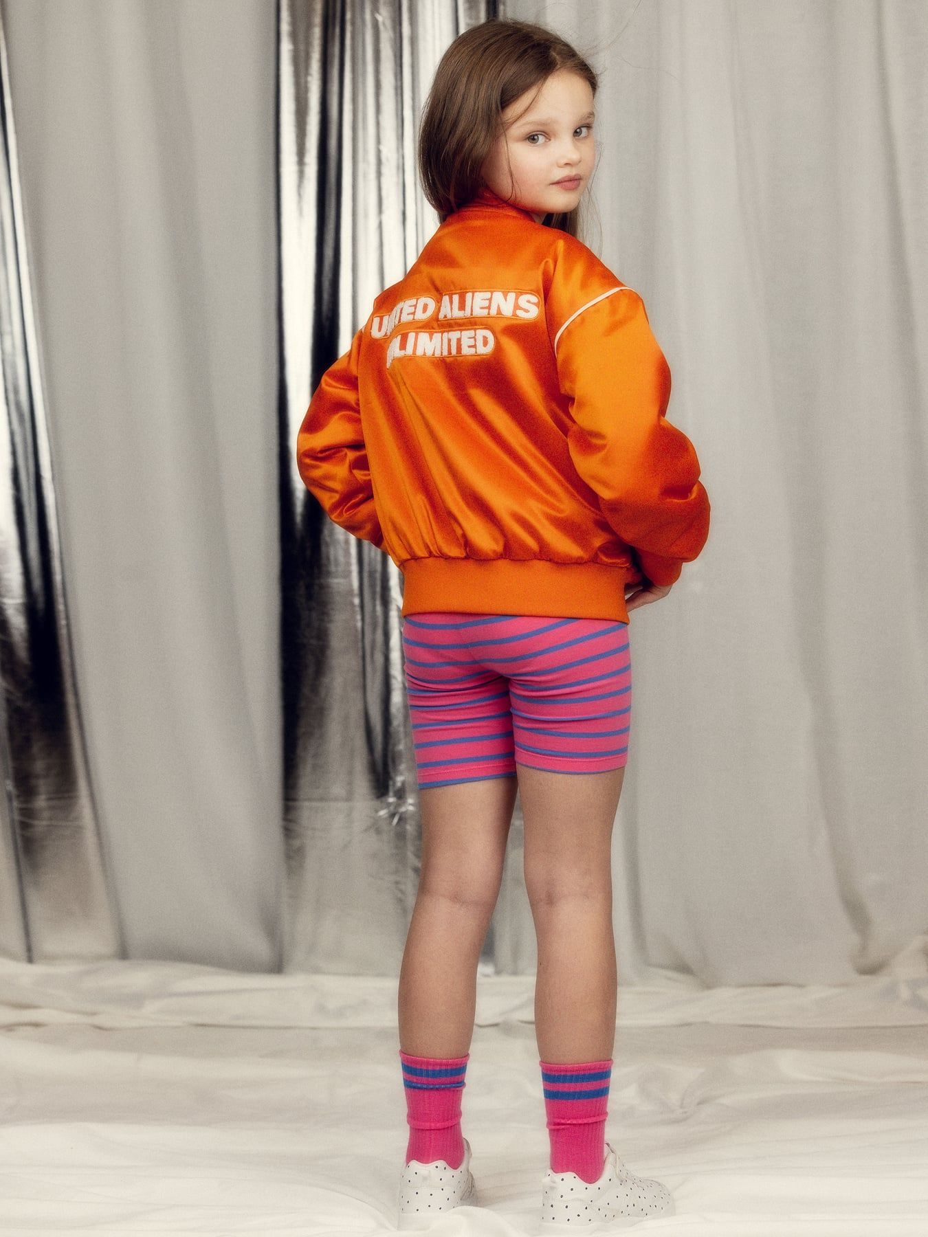 Child wearing an orange jacket with text, striped shorts, and colorful socks against a neutral background