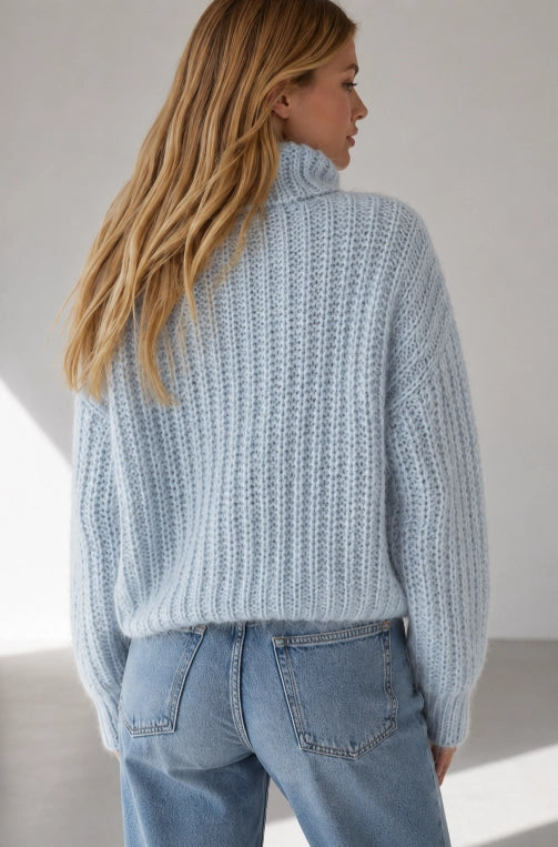 A woman shown from the back wearing a light blue ribbed knit turtleneck sweater and light blue jeans in a bright studio setting.
