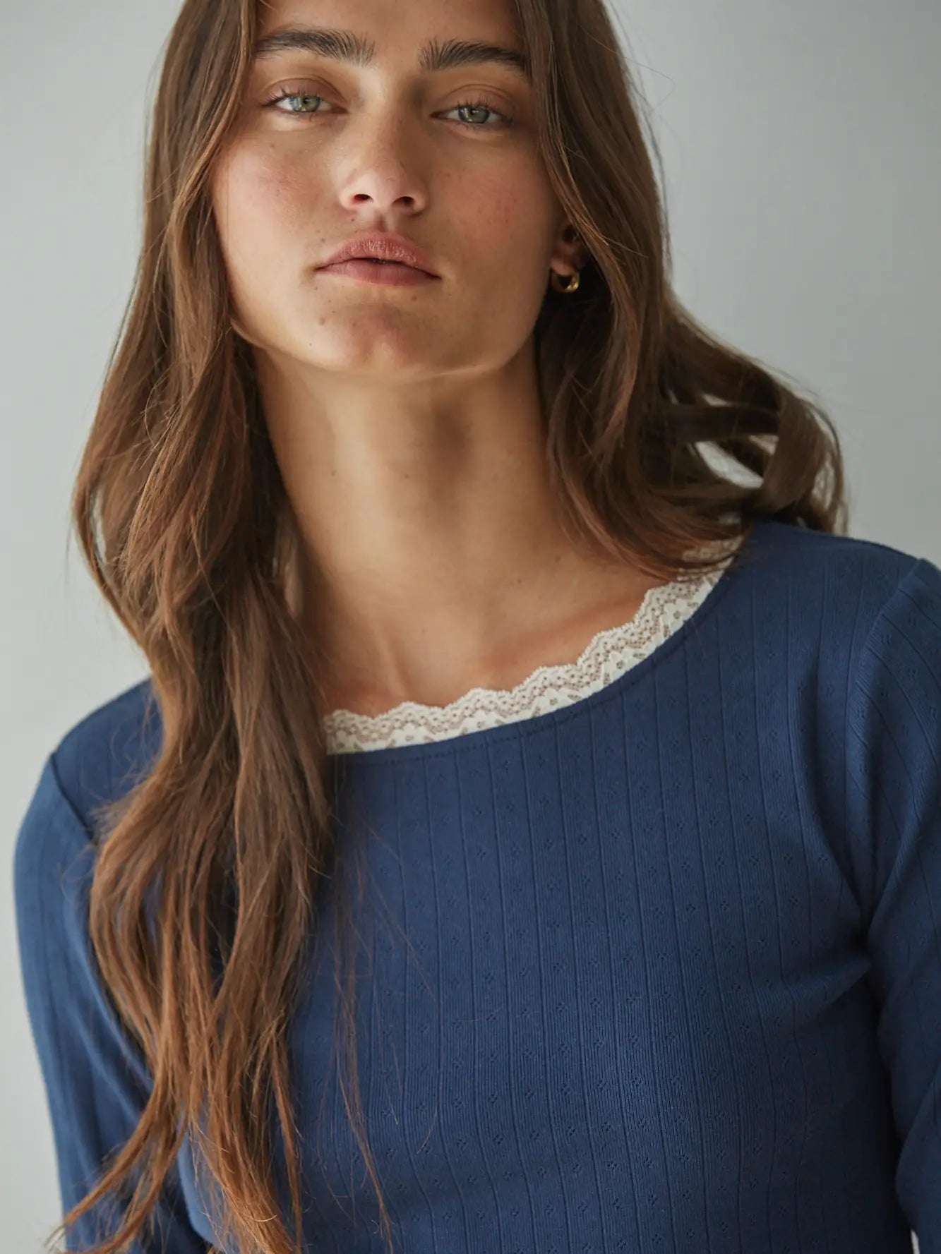 A close-up of a woman wearing a blue ribbed knit top with a white scalloped lace trim at the neckline, set against a plain light gray background.