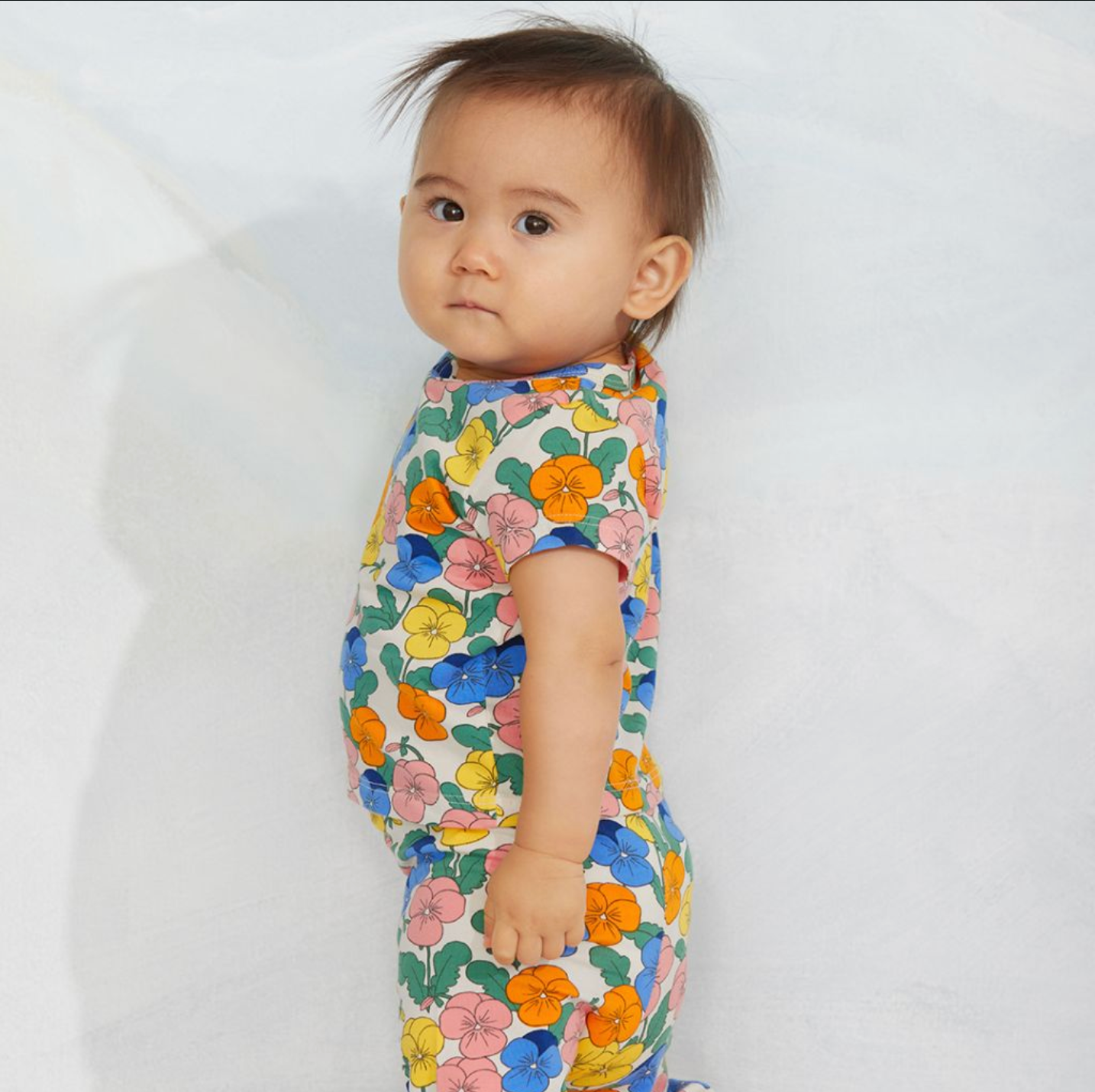 A baby wearing a short-sleeve one-piece romper with a colorful floral print, standing against a plain white background.