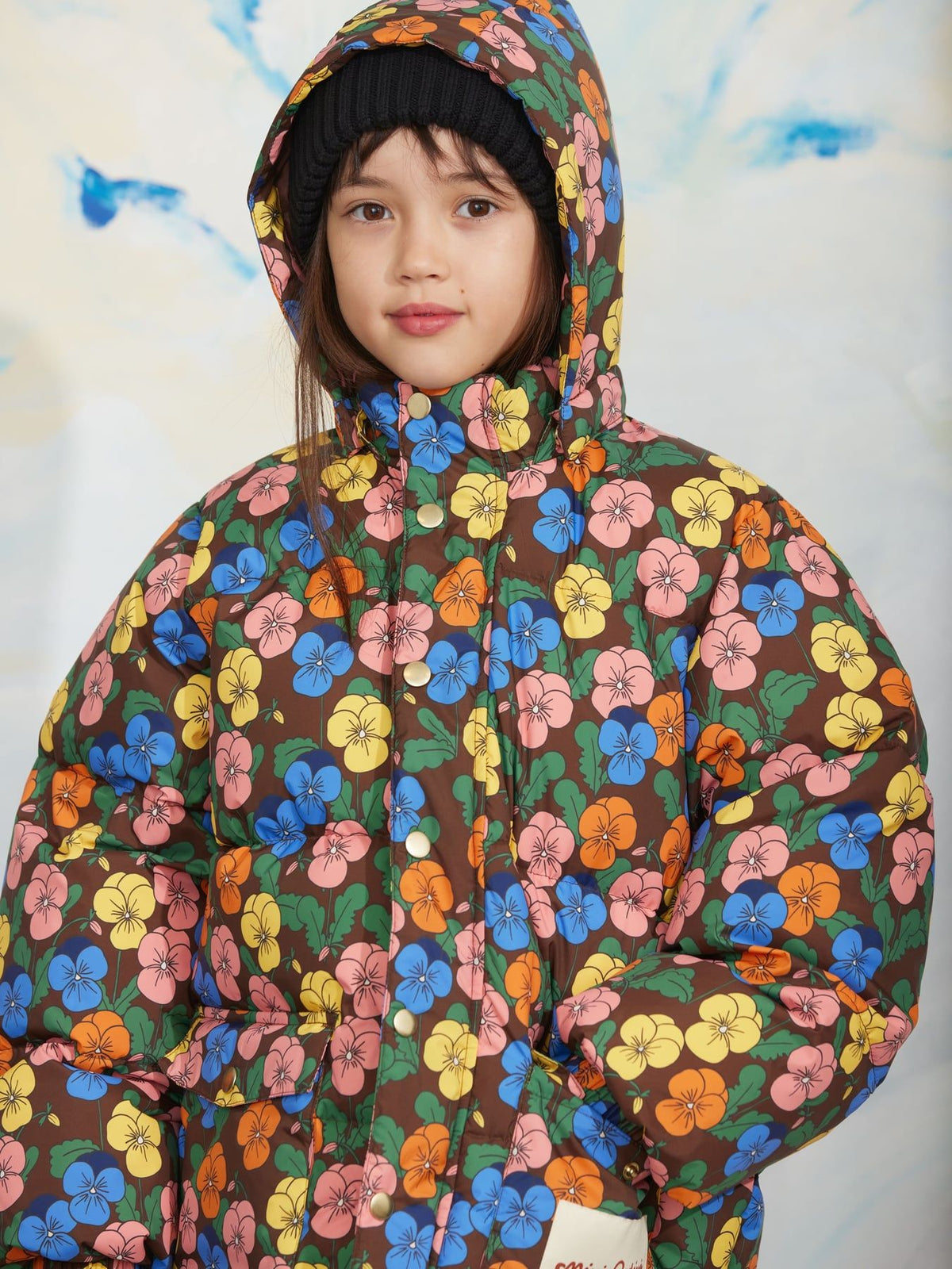 A child wearing a brown puffer jacket covered in a colorful floral print, paired with a black beanie, against a light background with blue abstract paint strokes.