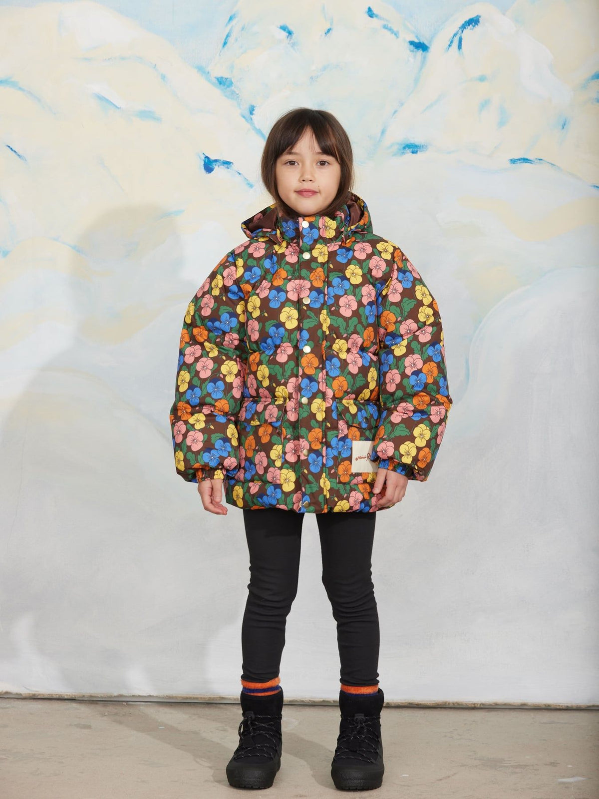 A child model wearing a colorful floral print puffer jacket, black pants, and black boots, standing in front of a stylized white and blue painted backdrop.