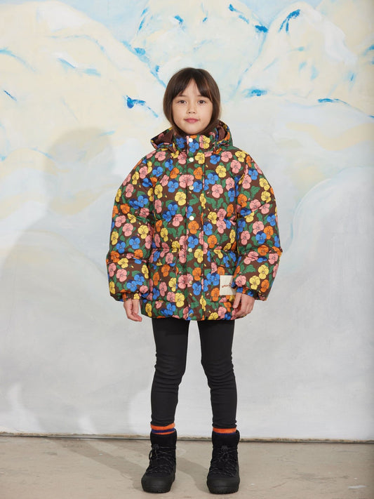 A child model wearing a colorful floral print puffer jacket, black pants, and black boots, standing in front of a stylized white and blue painted backdrop.