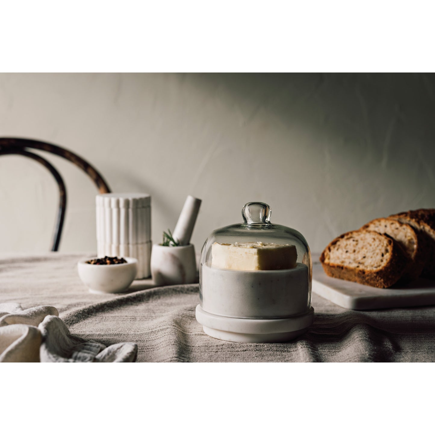 White ceramic container with glass lid on a table with bread and decorative items.