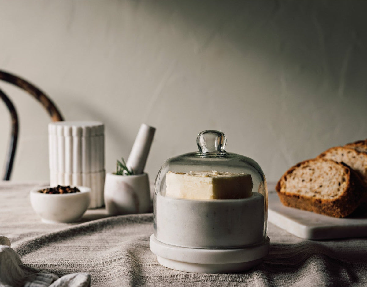 White ceramic container with glass lid on a table with bread and decorative items.