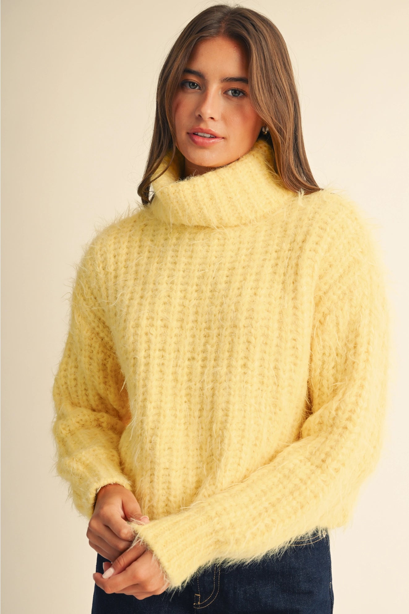 A woman wearing a fuzzy, textured pale yellow turtleneck sweater, paired with dark blue jeans, against a solid light beige background.