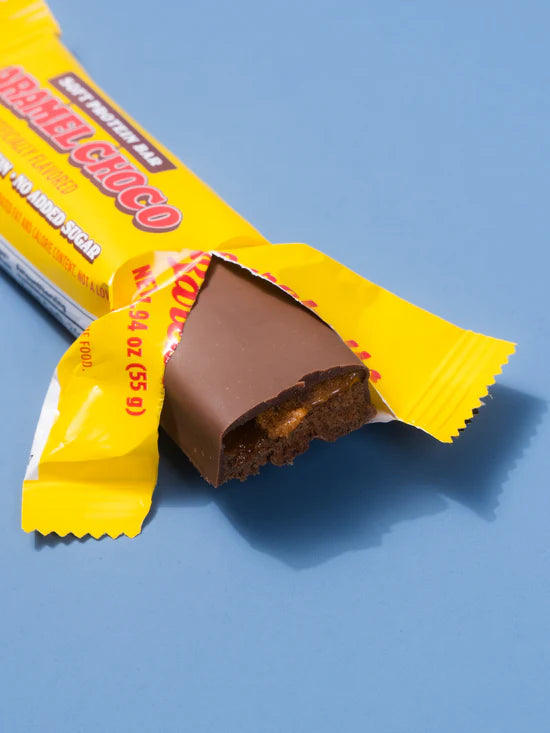 A partially unwrapped caramel chocolate cookie bar on a solid blue background. The bright yellow wrapper shows the text 'Caramel Choco' and 'Soft Pretender Bars', with a bite of the chocolate cookie bar exposed.