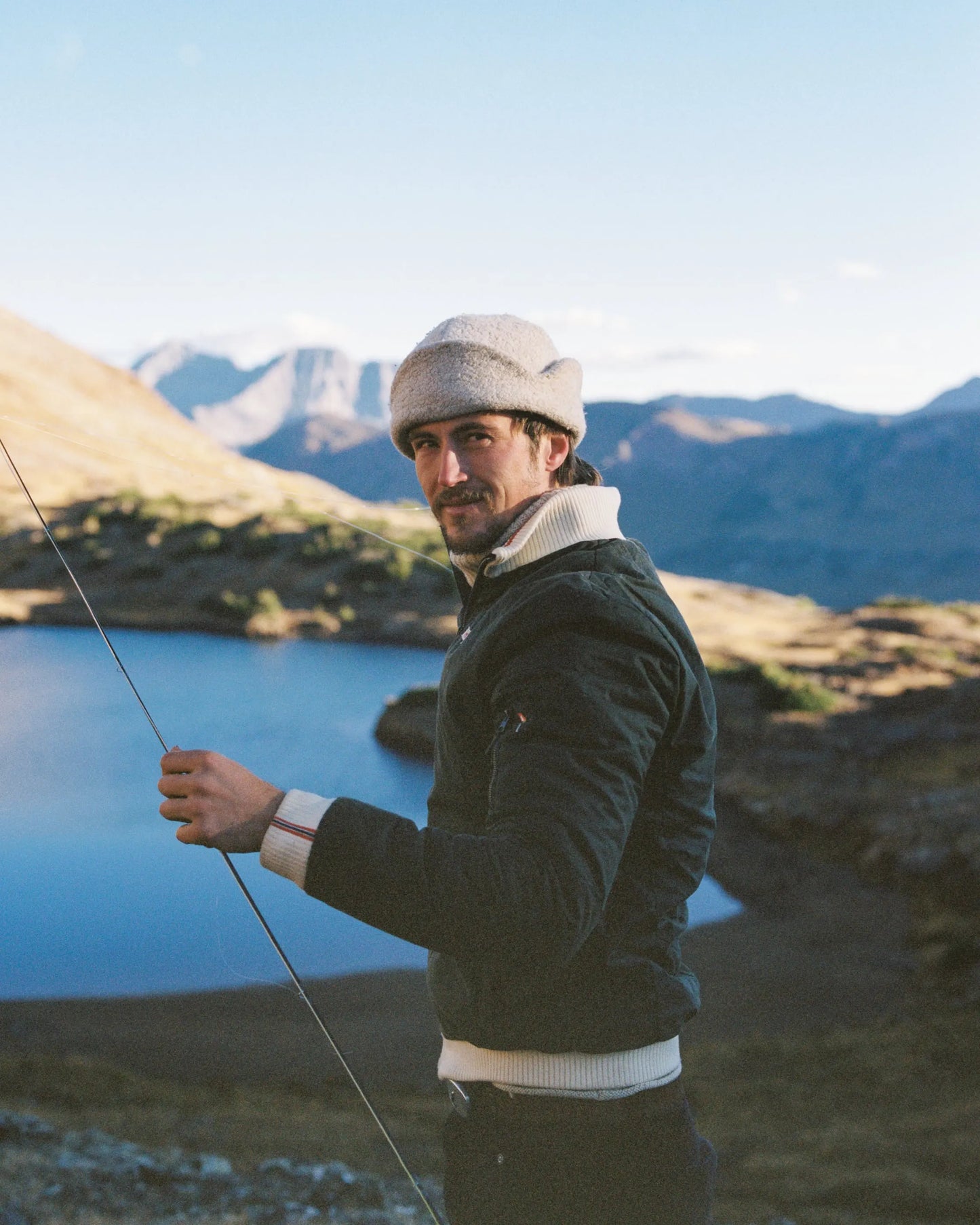 A man wearing a dark green waxed cotton jacket, white knit collar, and beige knit beanie stands outdoors near a blue lake with mountains in the background, holding a fishing rod.