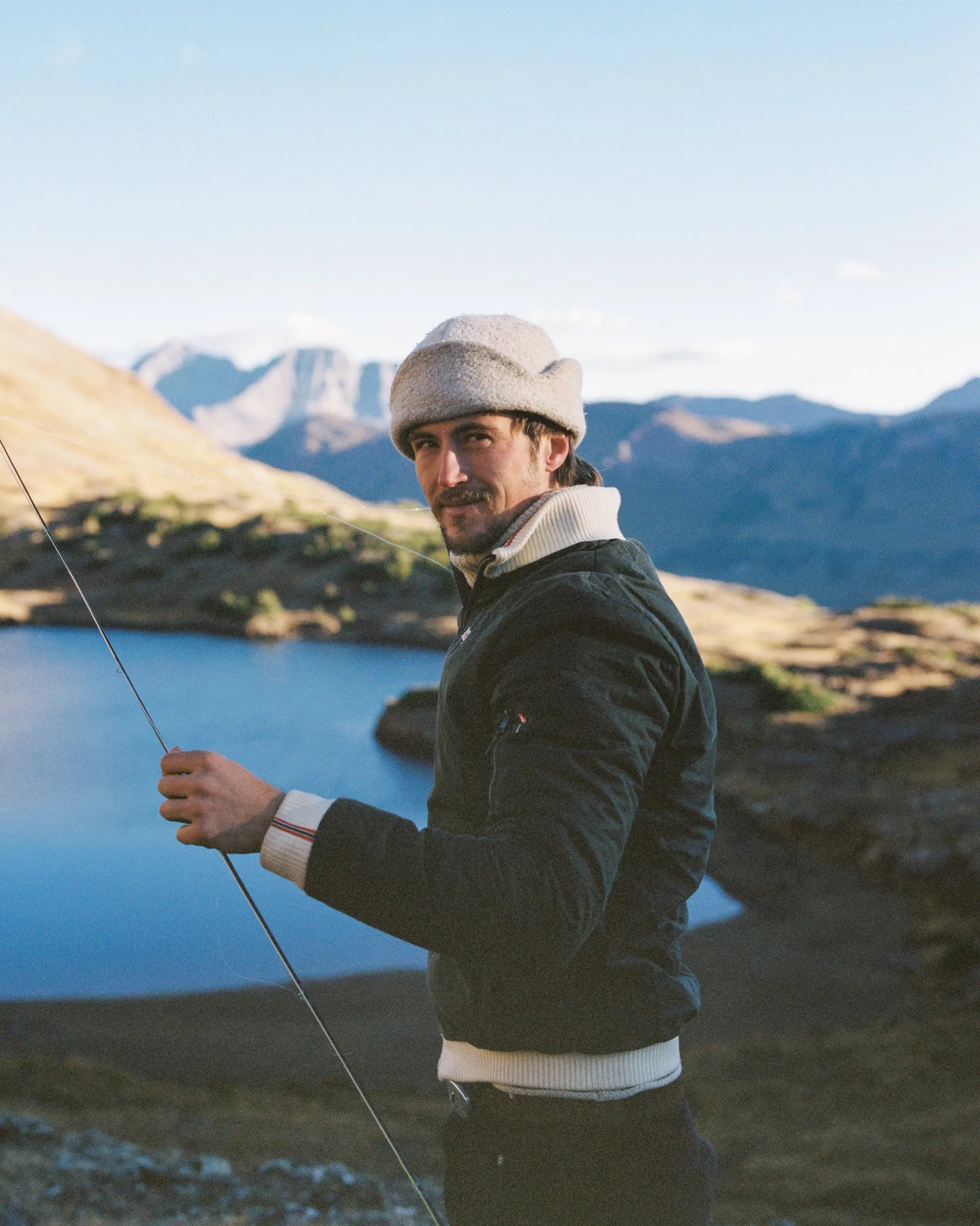 A man wearing a dark green waxed cotton jacket, white knit collar, and beige knit beanie stands outdoors near a blue lake with mountains in the background, holding a fishing rod.