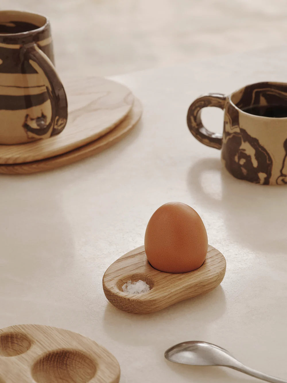 Wooden egg holder with an egg on a light surface with mugs and a spoon.