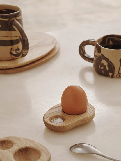Wooden egg holder with an egg on a light surface with mugs and a spoon.