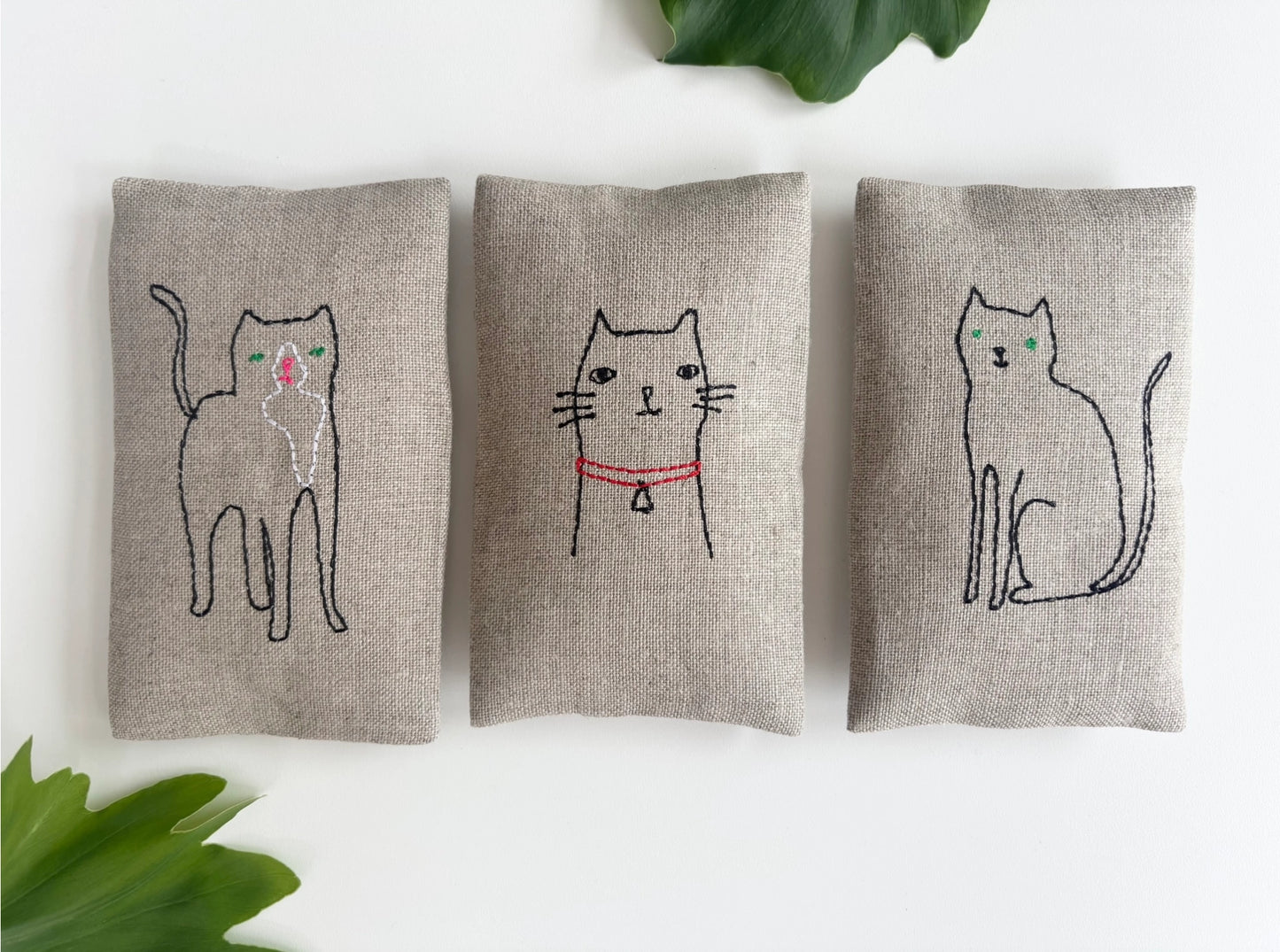 Three embroidered cat lavender sachet set on a white background with green leaves.