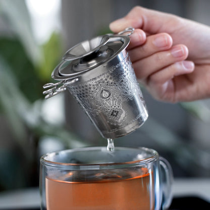 Person pouring tea into a glass mug using a metal tea infuser.