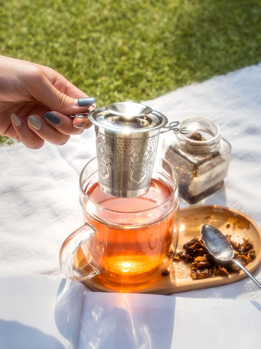 Tea being brewed in a glass teapot with a metal infuser on a white surface outdoors.