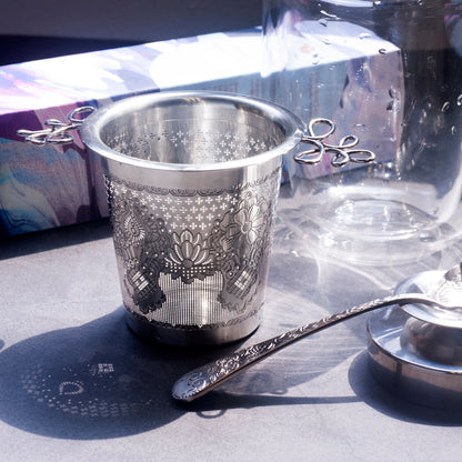 Silver tea strainer with intricate designs on a reflective surface