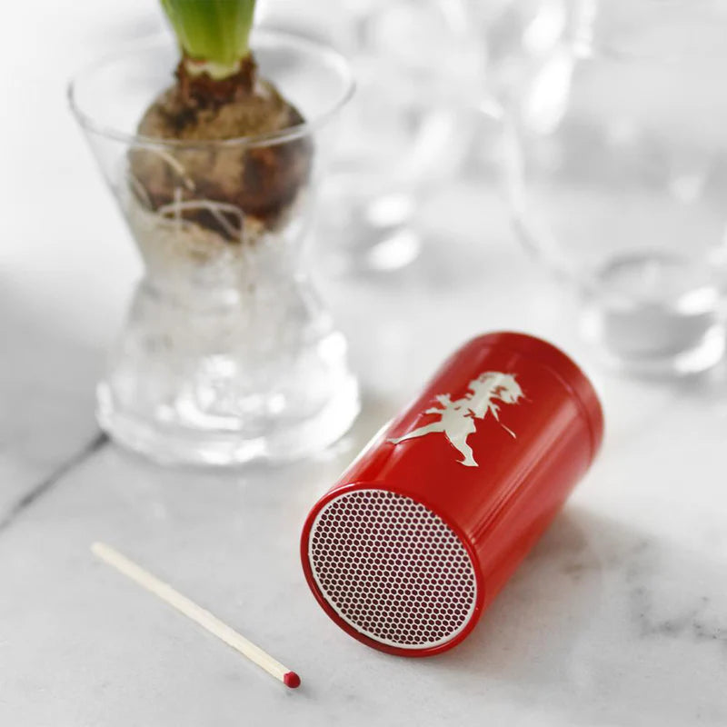 Red cylindrical matchbox with white design on a marble surface with a plant in the background